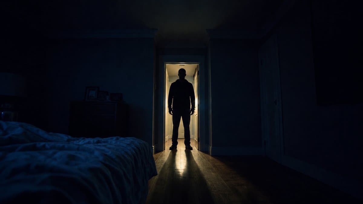 Dunkle Silhouette einer Person im Türrahmen eines nächtlichen Schlafzimmers, ein langer Lichtschatten fällt auf den Boden