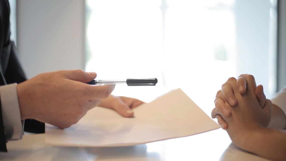 Ein Mann im Anzug reicht einer Person am Tisch einen Stift über ein Vertragsdokument, die Hände der Person sind gefaltet