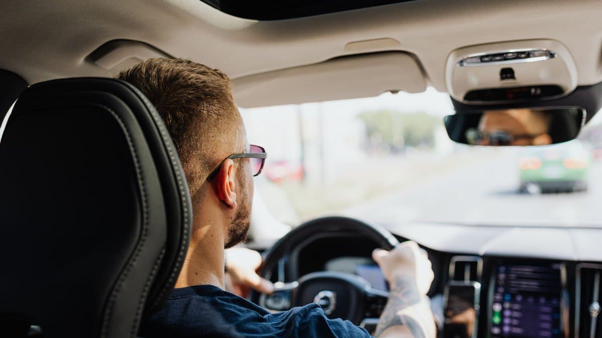 Mann am Steuer eines Autos, über die Schulter von der Rücksitzbank aus fotografiert, Blick durch die Windschutzscheibe auf die Straße
