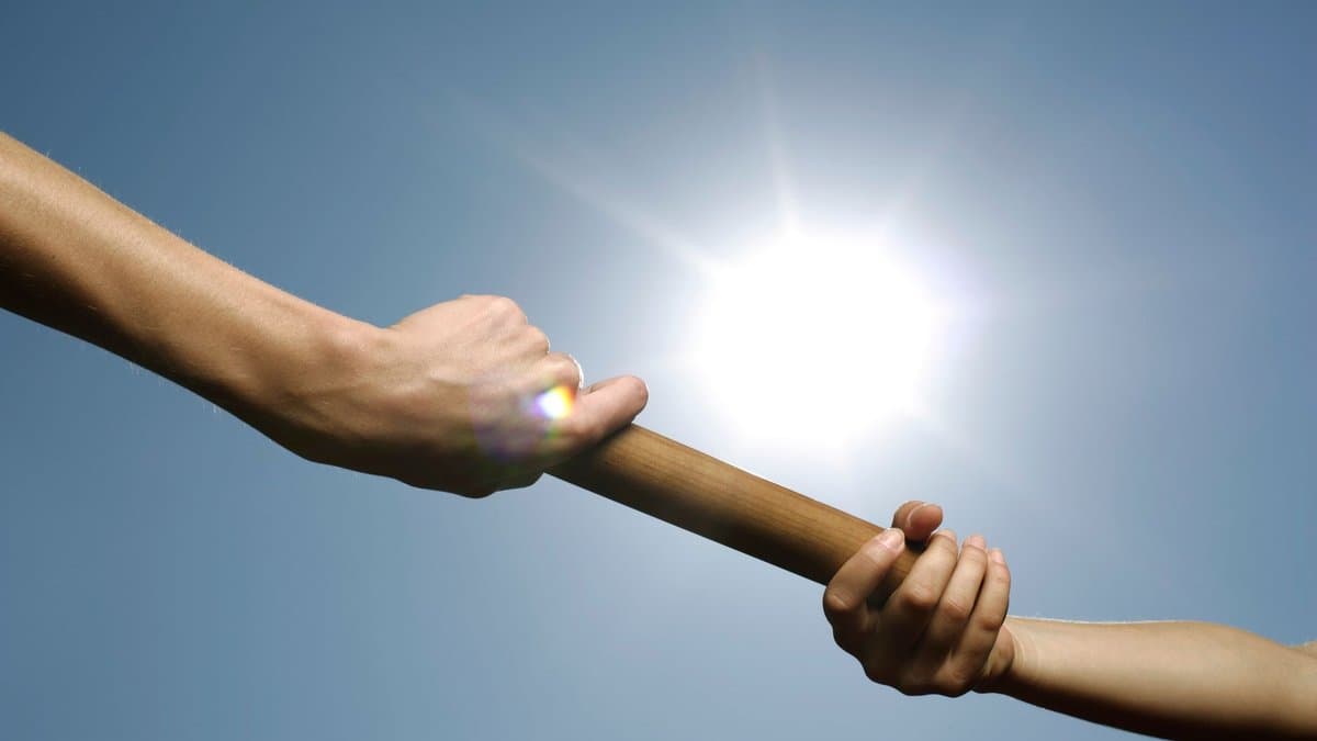 Zwei Hände übergeben einen Staffelstab vor blauem Himmel, die eine Hand gibt ab, die andere greift zu