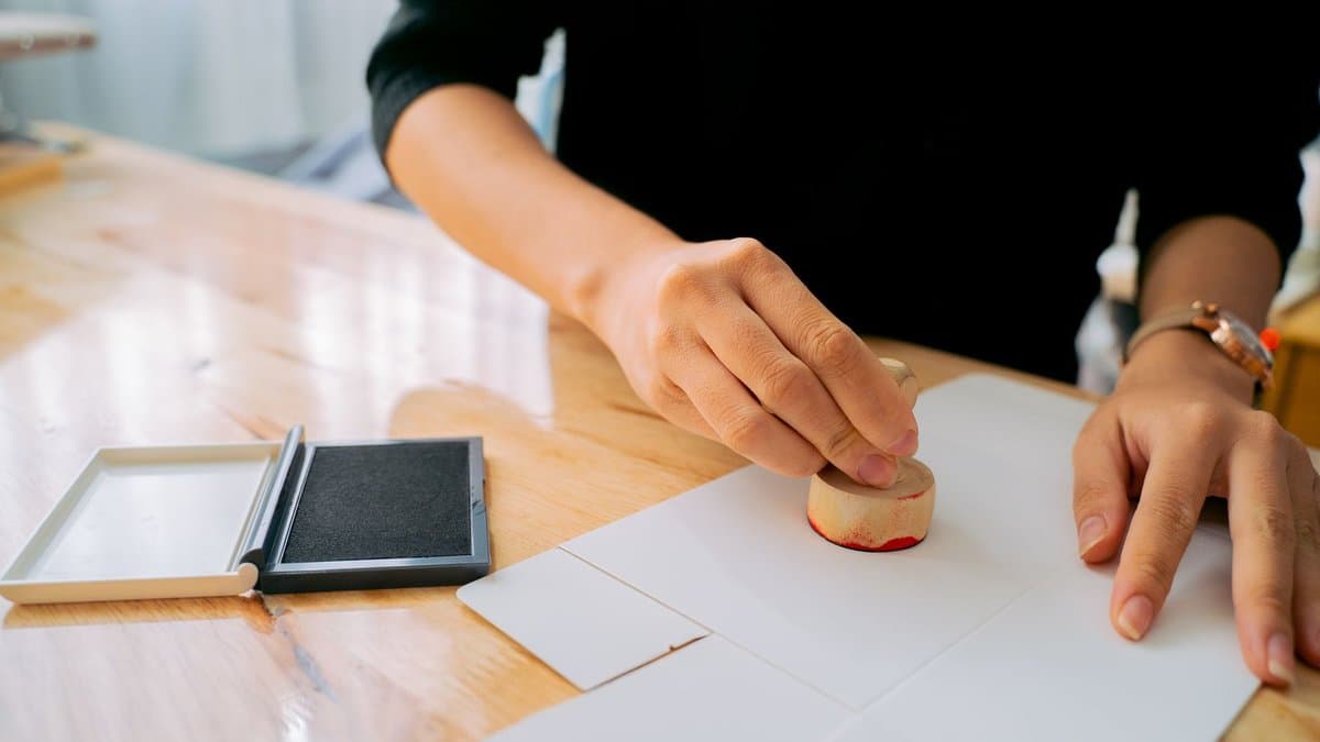 Hand setzt einen Holzstempel auf ein Blatt Papier auf einem hellen Holzschreibtisch, daneben ein aufgeklapptes Stempelkissen