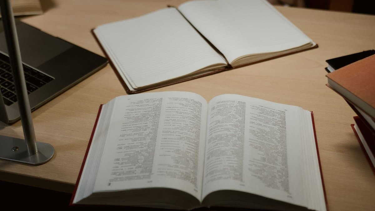 Aufgeschlagenes dickes Gesetzbuch mit rotem Einband auf einem Schreibtisch, daneben Notizbuch, Laptop und weitere Bände