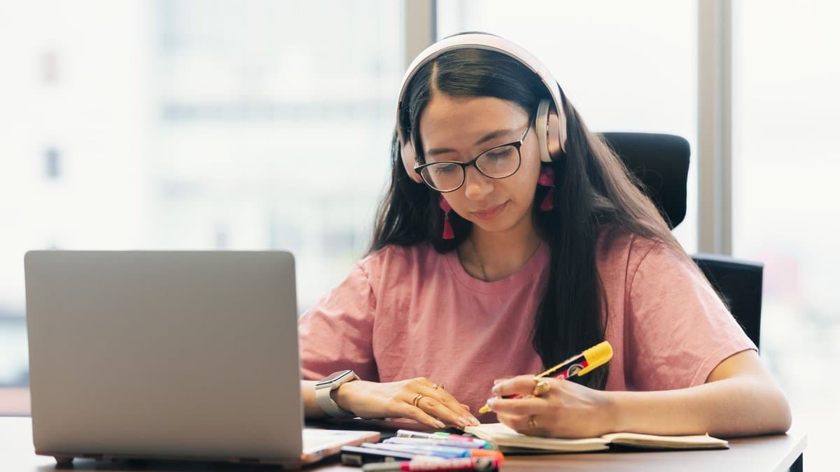 Studentin mit Kopfhörern sitzt vor einem Laptop und schreibt in einen Notizblock