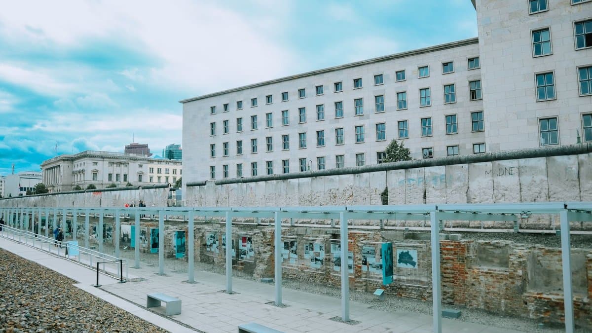 Erhaltenes Teilstück der Berliner Mauer entlang eines gepflasterten Wegs, dahinter ein großes Verwaltungsgebäude