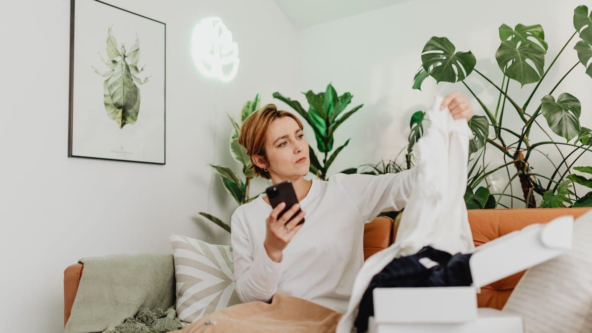 Frau sitzt auf dem Sofa mit dem Smartphone in der Hand und prüft ein bestelltes Kleidungsstück, daneben Versandkartons