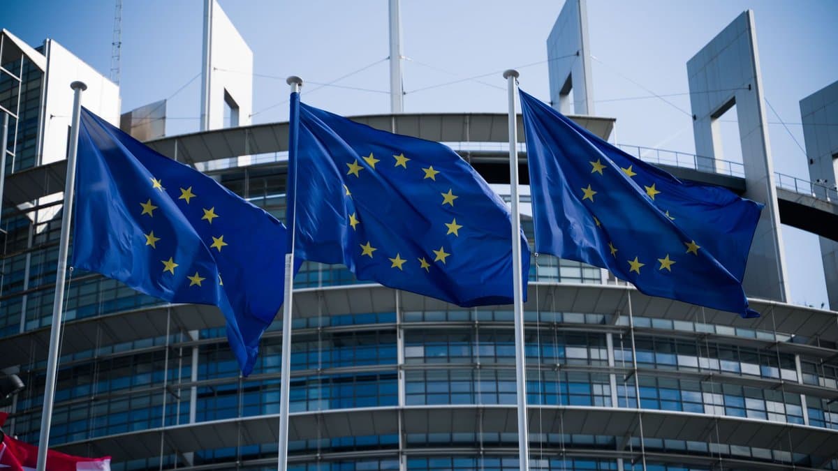 Drei Europaflaggen wehen vor der gläsernen Fassade eines EU-Parlamentsgebäudes unter blauem Himmel