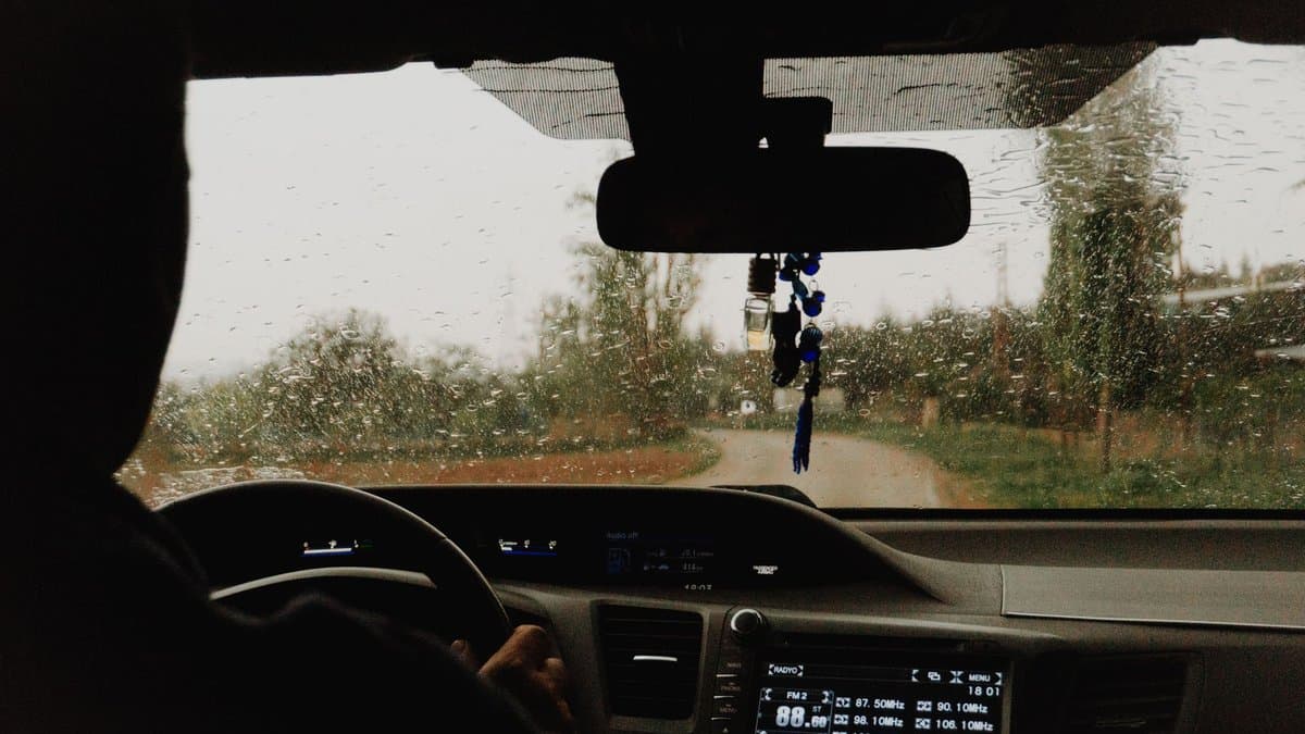 Blick durch die regennasse Windschutzscheibe auf eine schmale Landstraße, eine Hand am Lenkrad