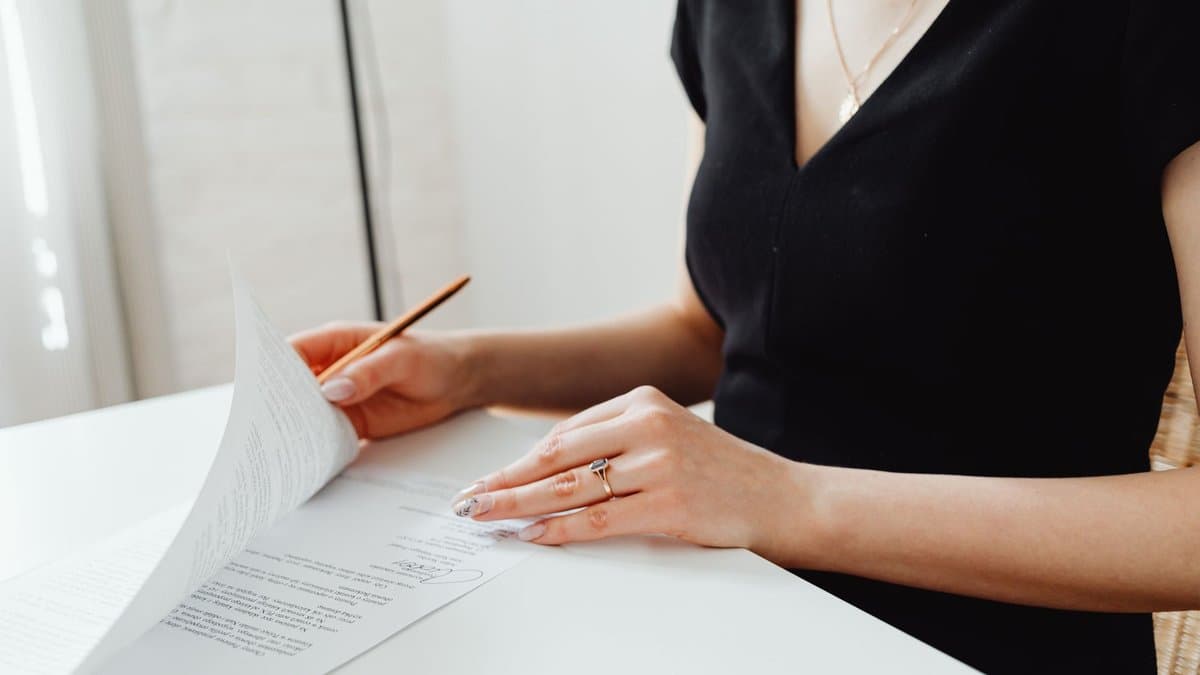 Person am Schreibtisch hebt eine Vertragsseite an und liest sie, Stift in der Hand