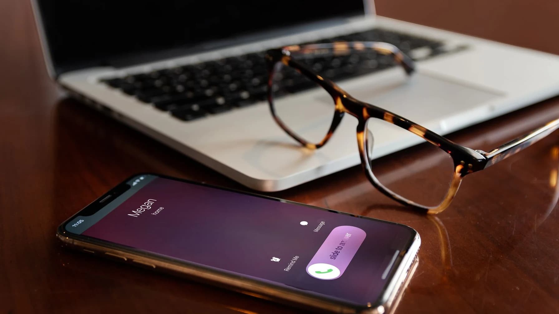 Ein Smartphone mit eingehendem Anruf liegt auf einem Schreibtisch neben einem Laptop und einer Brille