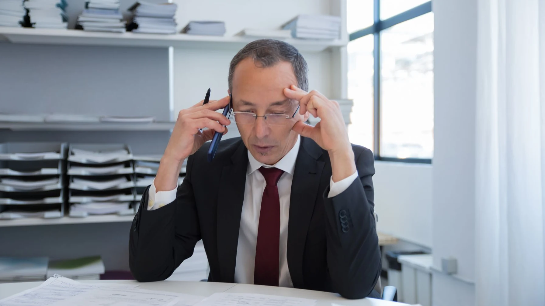 Mann im Anzug mit Brille telefoniert am Schreibtisch, vor ihm liegen Dokumente.