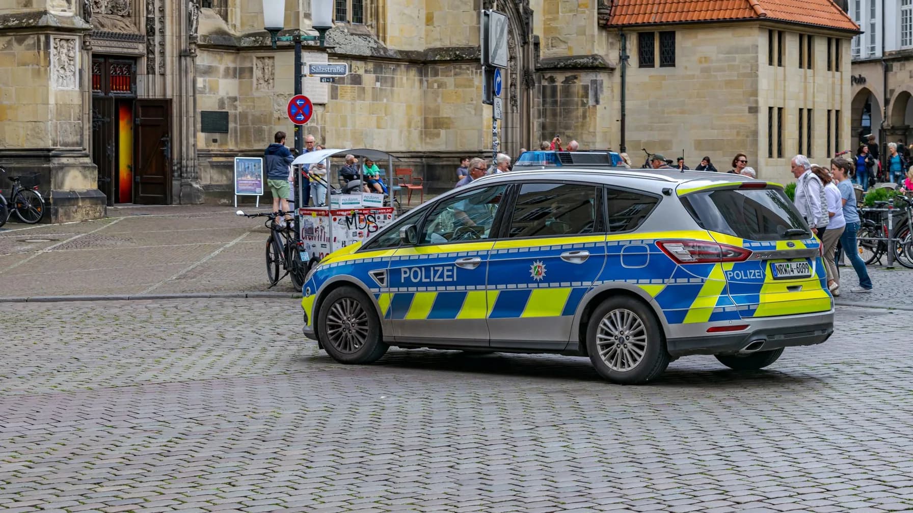 Streifenwagen der Polizei auf einem historischen Stadtplatz, im Hintergrund Passanten und Radfahrer