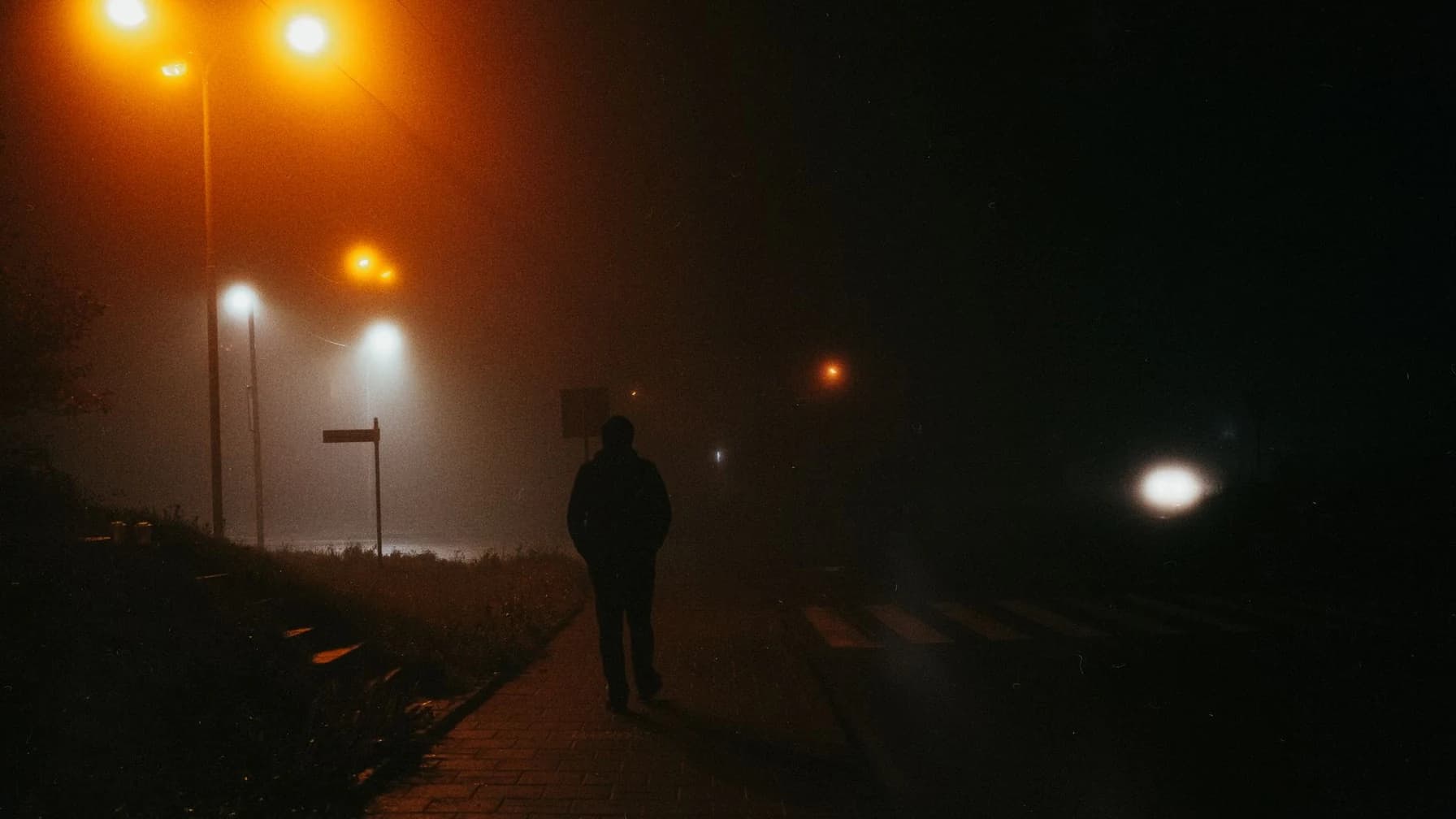 Mann geht nachts allein über einen nassen, nebligen Gehweg an leuchtenden Straßenlaternen vorbei, von hinten gesehen