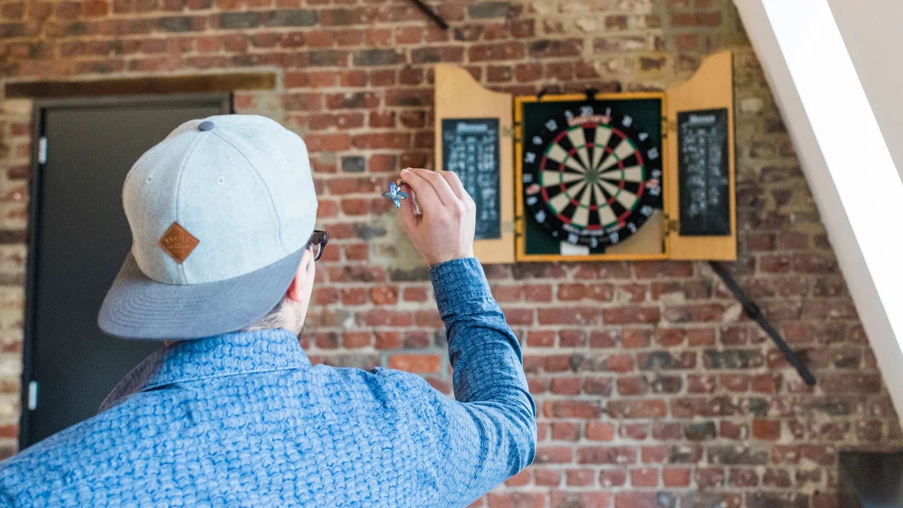 Ein Mann von hinten wirft einen Dartpfeil und zielt konzentriert auf eine Dartscheibe an einer Backsteinwand