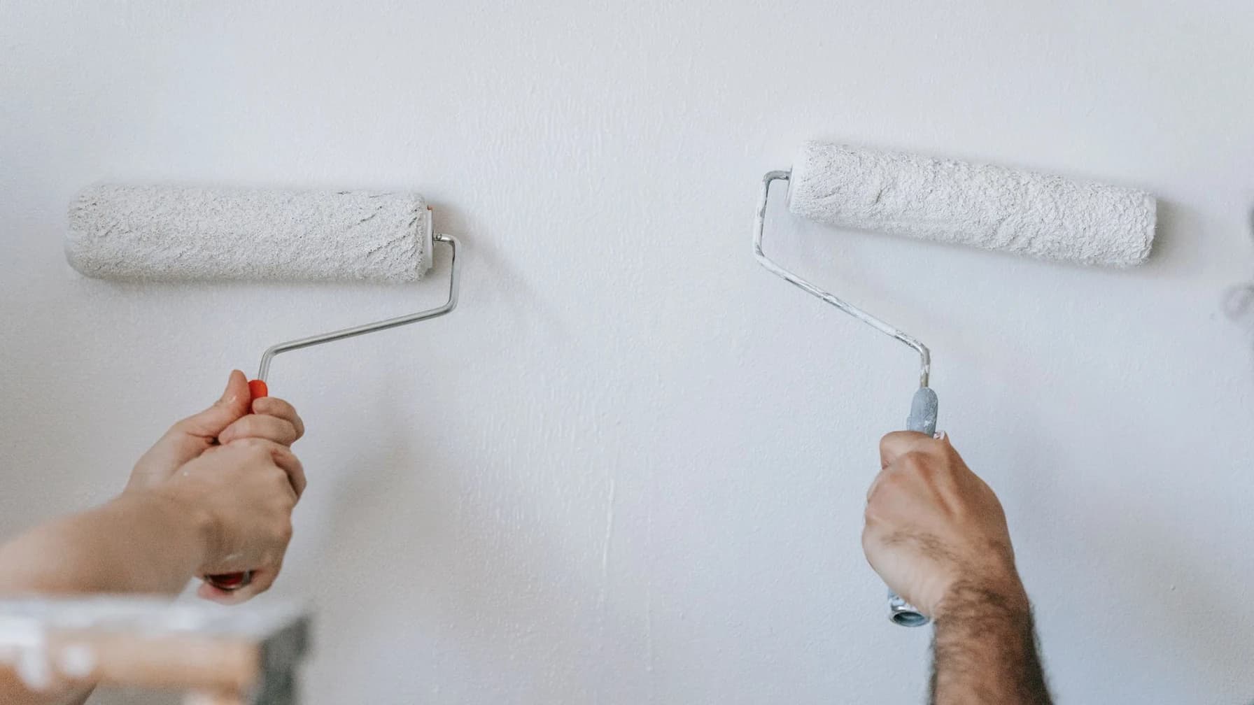 Zwei Hände streichen mit weißen Farbrollen eine helle Wand
