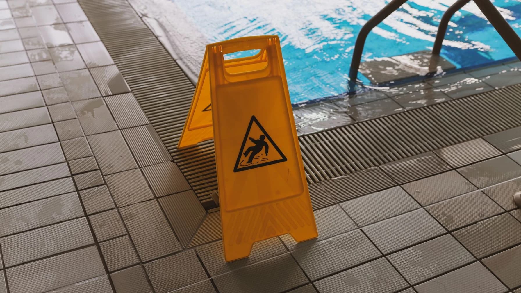 Gelbes Warnschild mit Rutschgefahr-Symbol auf nassen Fliesen neben einem Schwimmbecken